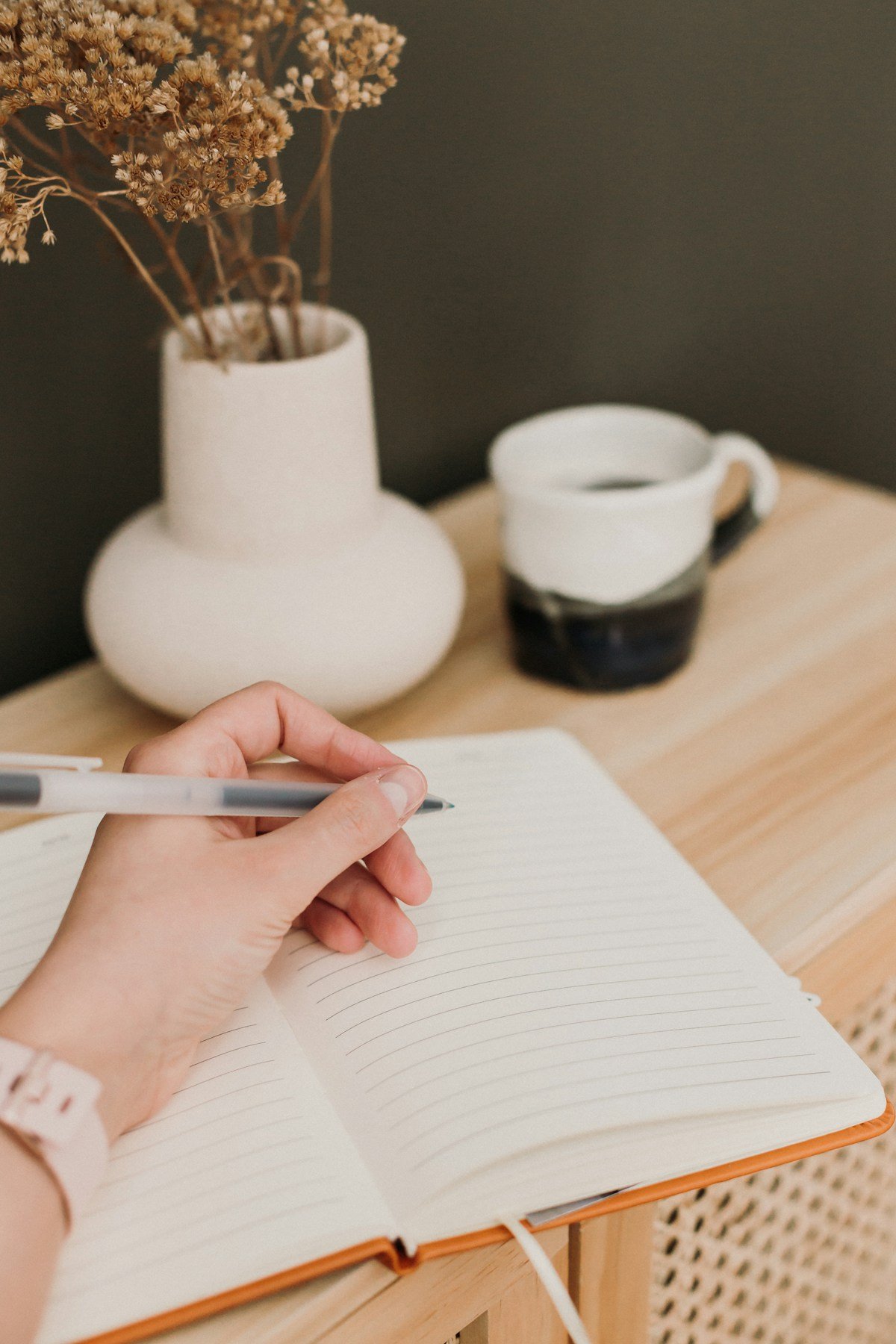 Manos escribiendo en un cuaderno, junto a una taza y un florero con flores secas